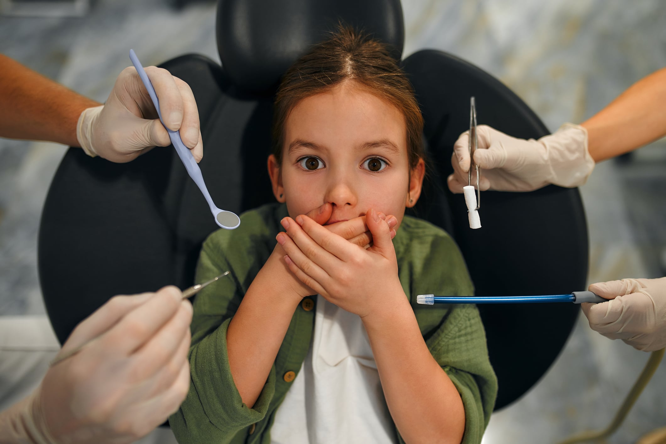 Pleasant little girl lying in a dentist chair and covering her mouth, being afraid of the checkup at the dentists.